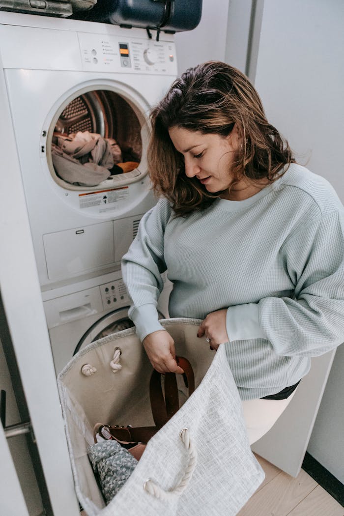 Young woman with long hair in casual clothes putting clothes from bag into washing machine in laundry room